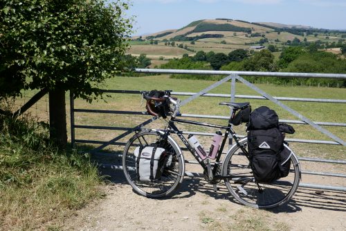 A loaded touring bike leaning against a gate, with fields, trees and a hill in the background