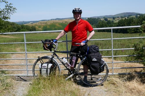 A man in a red short-sleeved top standing behind a loaded touring bike. Fields and hills in the background.