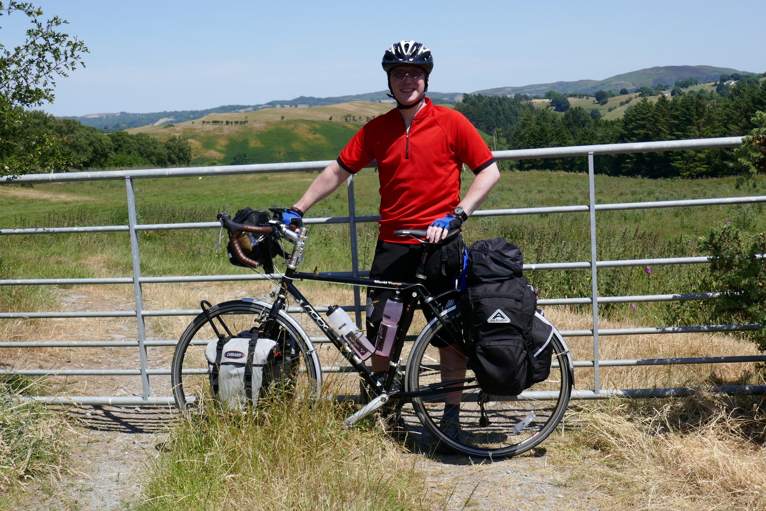A man in a red short-sleeved top standing behind a loaded touring bike. Fields and hills in the background.
