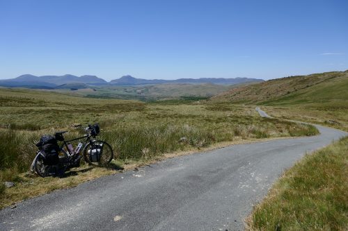 A loaded touring bike stands beside a narrow, winding road through miles of bare green hills, with mountains in the distance.
