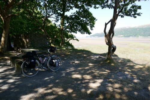 A loaded touring bike rests again a picnic table in the shade of twisted trees. Tree-covered hills in the background.
