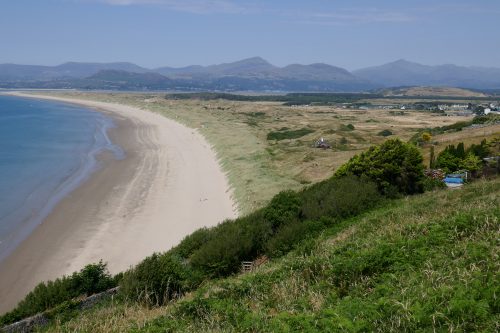 Golden sandy beach curving into the distance, between blue sea and grass covered land. A mountain range in the background.