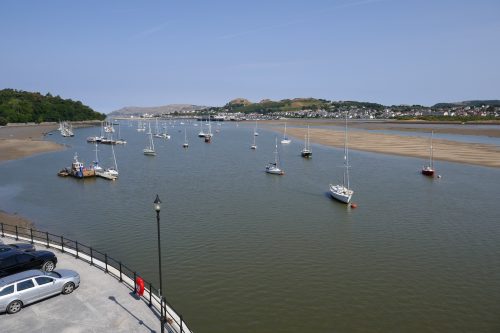 Boats in a harbour, with sandbanks. In the background are houses at the foot of a green hillside.