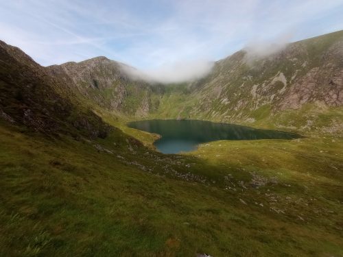 A small blue lake, surrounded by grass, sitting beneath mountain peaks over which white clouds are rolling.
