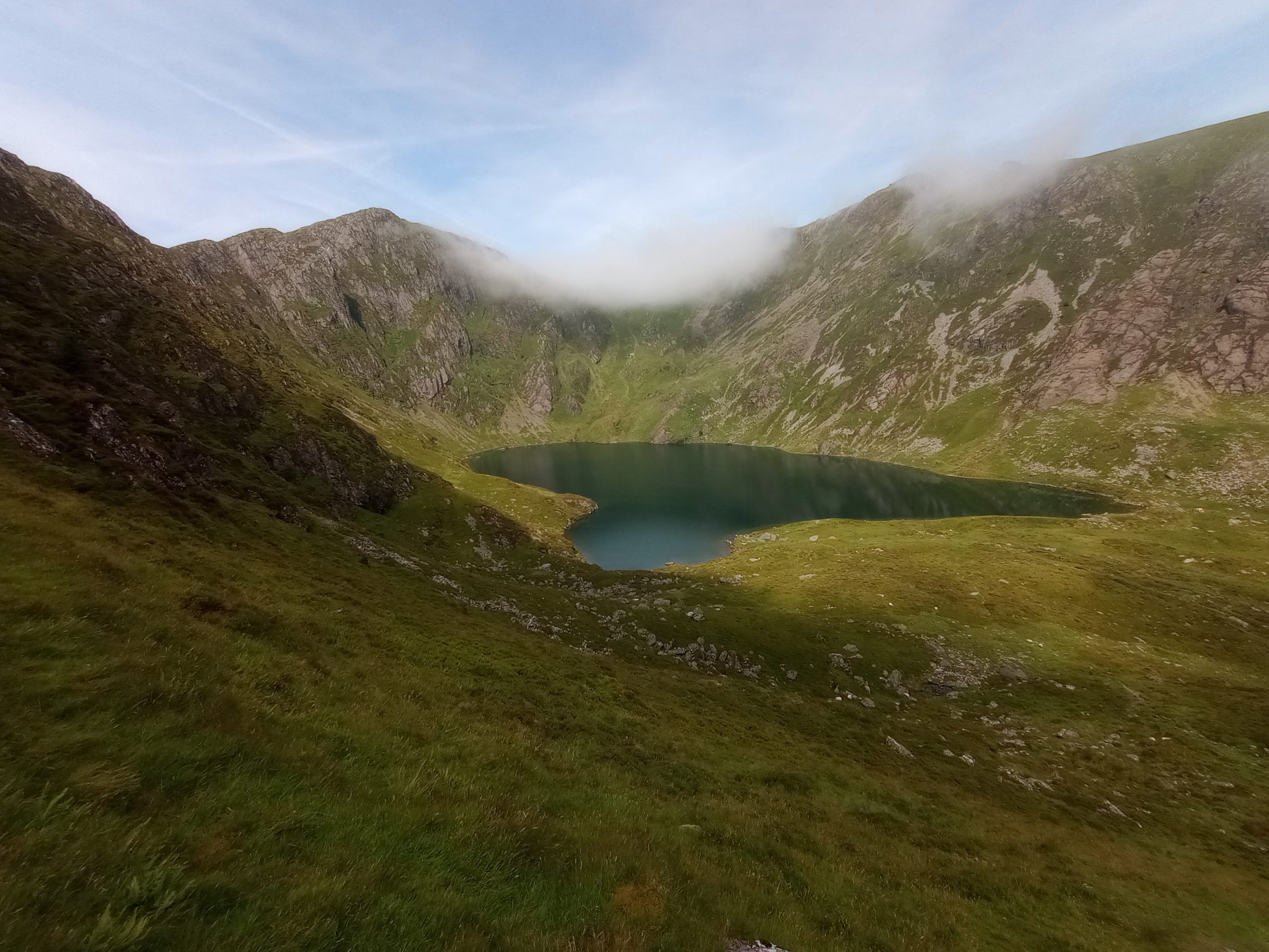 A small blue lake, surrounded by grass, sitting beneath mountain peaks over which white clouds are rolling.