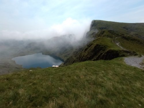 A small blue lake beneath a rocky mountain peak, with clouds on the shoulder. A couple of sheep are grazing.
