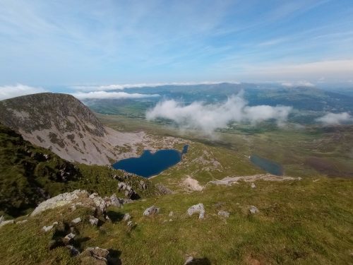 A small blue lake beneath a rocky mountain peak. Strings of cloud hover over the landscape into the distance.